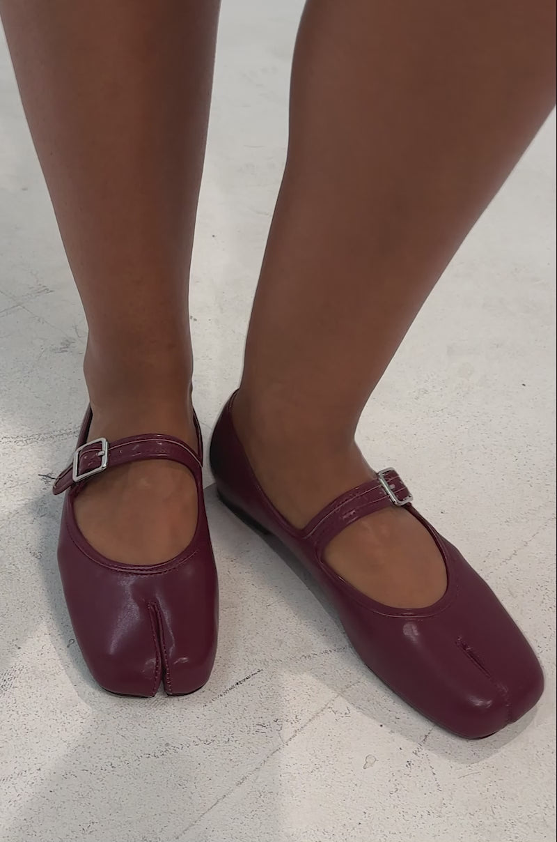 A video of a model wearing the AKIRA GOLD SANG BURGUNDY PU SPLIT TOE FLAT, featuring buckle straps and a comfort cushion insole.