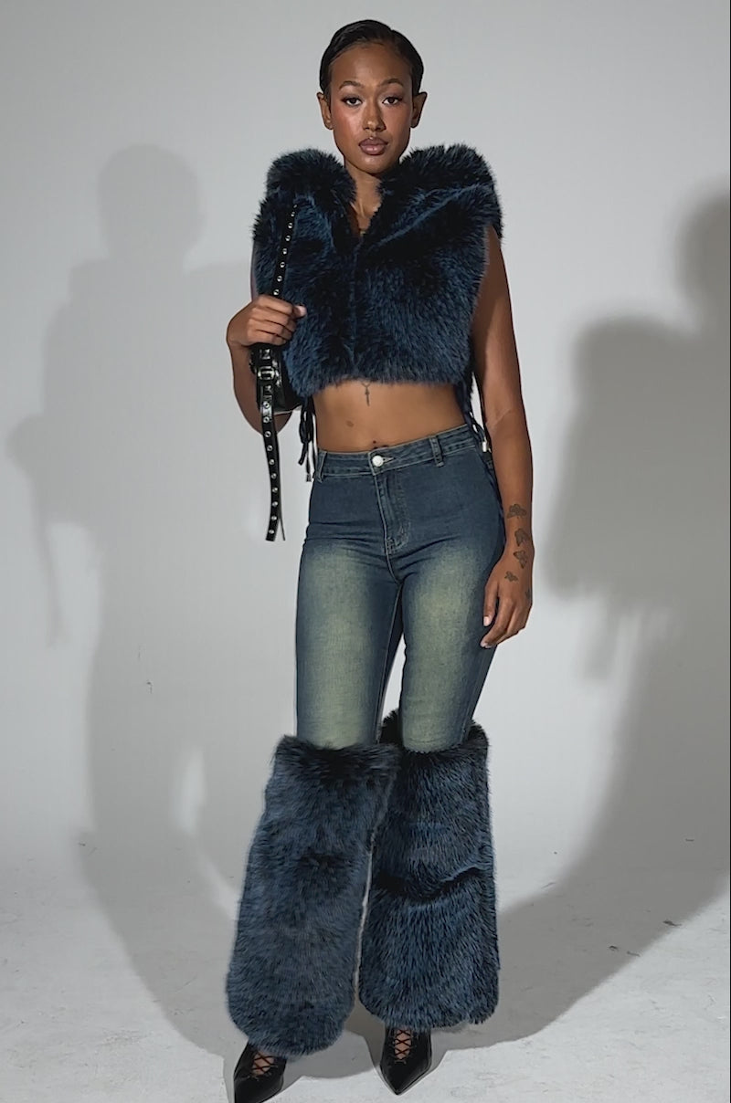 A video of a woman posing in the ALL A DREAM DENIM PANT WITH FUR CUFFS, styled with blue faux fur leg warmers, a blue faux fur cropped vest, black heels, and a black purse against a light background.