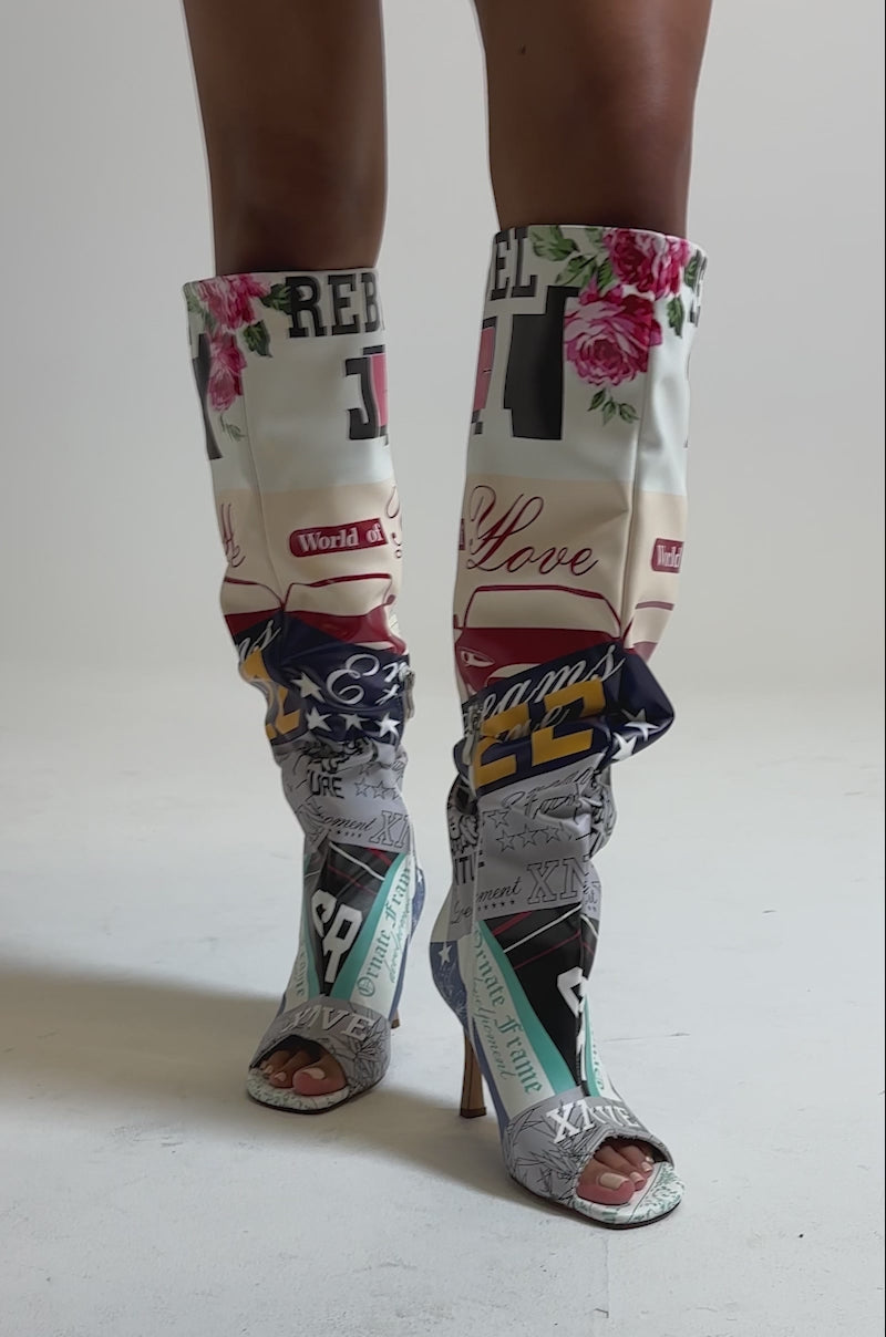 A video of a model wearing the AZALEA WANG JALISA MULTI NOVELTY PRINTED OPEN TOE BOOT, which features colorful patterns, bold text, florals, American flag motifs, and graphics on a light background. 