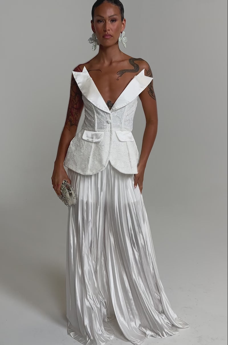 A video of a model wearing the CALLED OUT LACE CORSET and a sheer, flowing skirt, the person stands against a plain light background, holding a small embellished clutch.