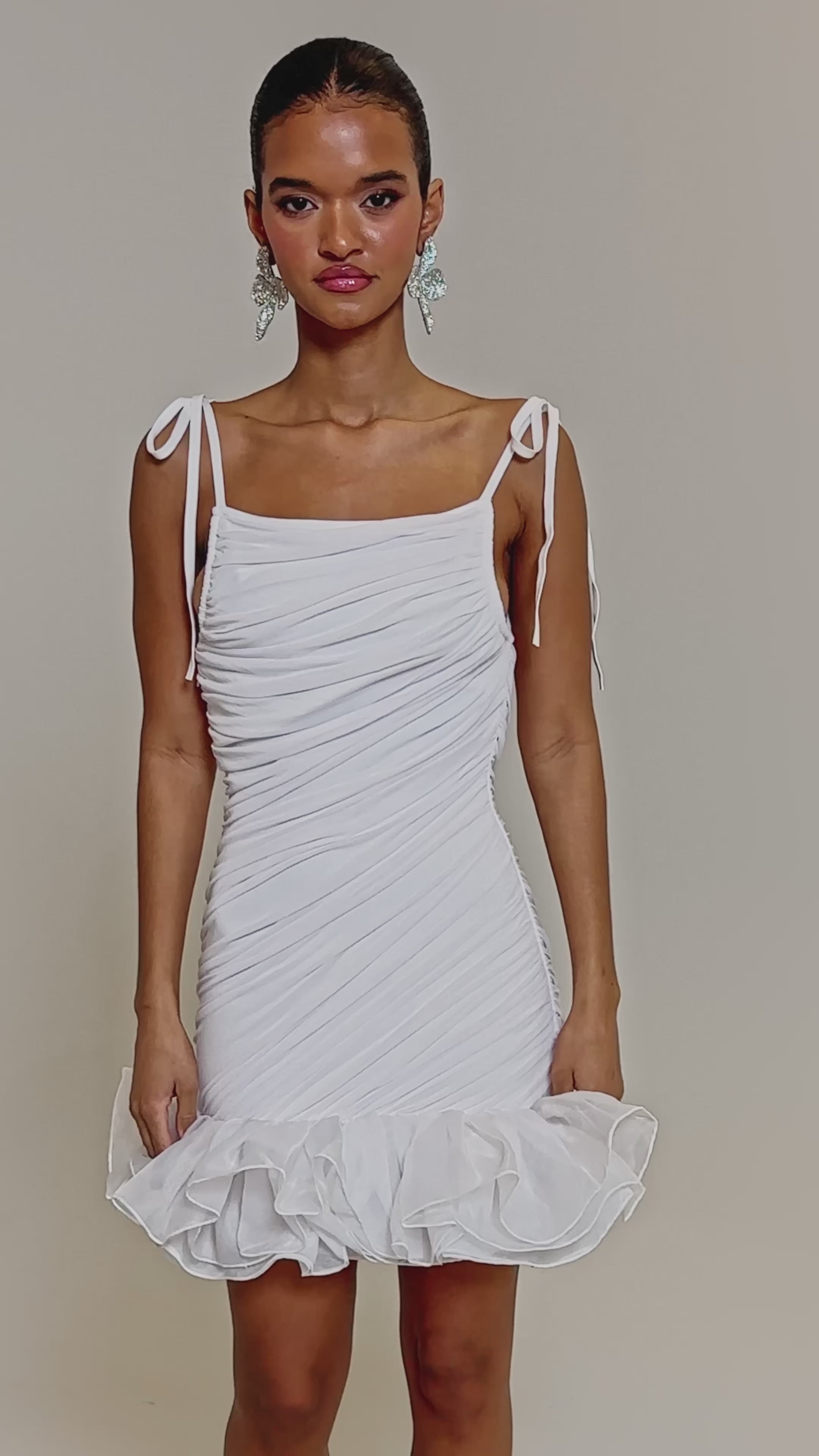 Video of woman modeling the PRINCESS PEPLUM MINI DRESS IN WHITE, featuring a fitted ruched mesh design with shoulder tie straps and an organza ruffle hemline. Her hair is slicked back and she accessorizes with large floral statement earrings.