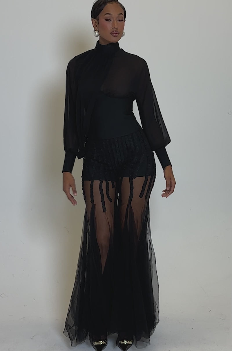 Video of woman modeling the FIRST THING FIRST BLOUSE, a sheer black top with billowy sleeves, paired with high-waisted textured black pants, accessorized with pearl earrings.