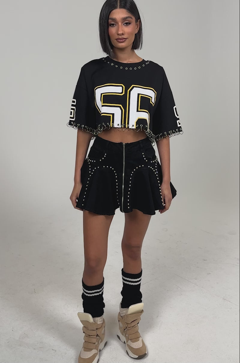 Video of woman modeling the LUCKY ONE DENIM MINI SKIRT with stud embellishments, paired with a black cropped top, black leg warmers, and beige chunky sneakers. She has shoulder-length hair and stands against a plain light background.