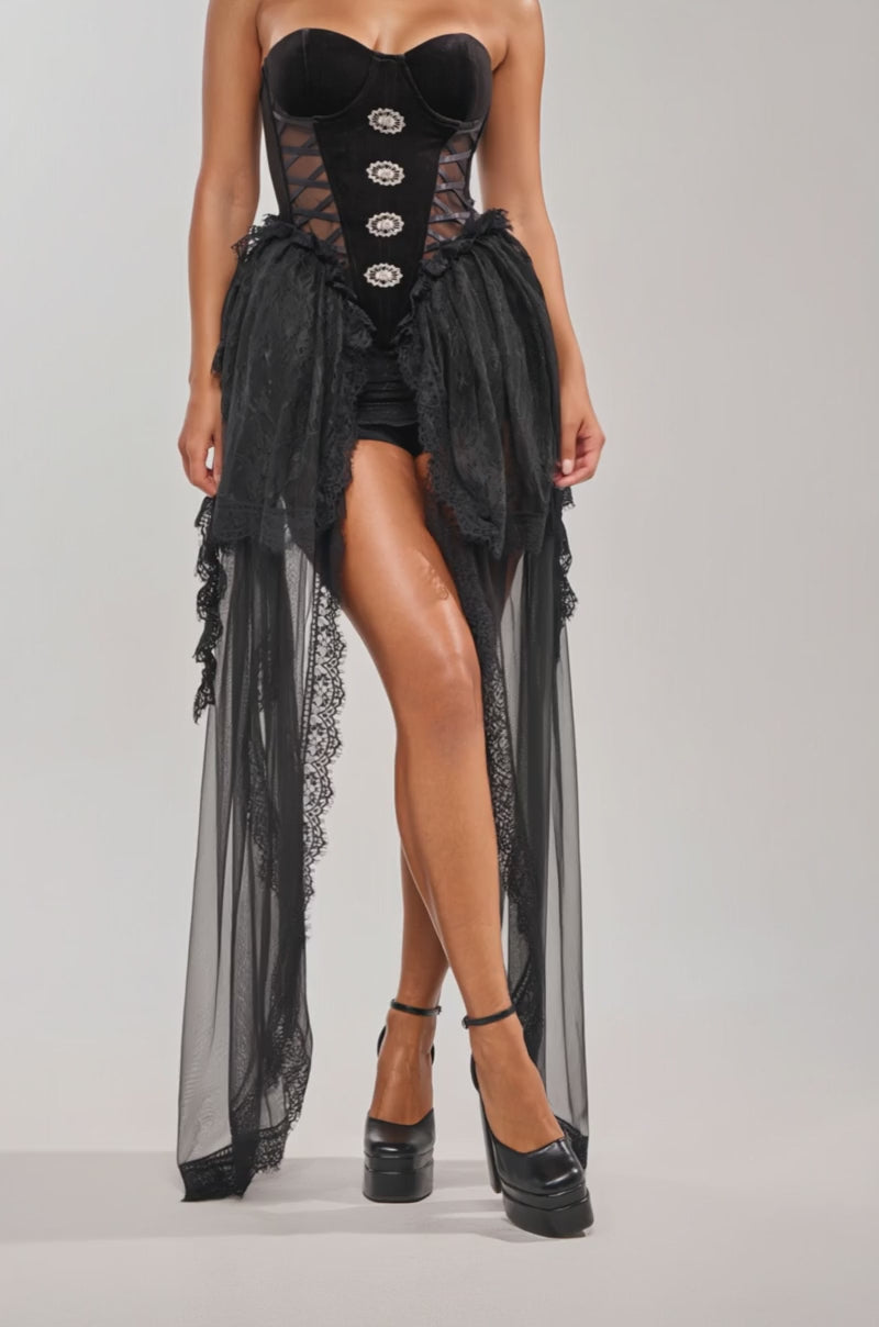 Video of woman modeling the SHE IS DRAMA CORSET, a black gothic dress with a sweetheart neckline, sheer lace-trimmed overlay, and shorts, styled with black platform heels and statement earrings against a plain light background.