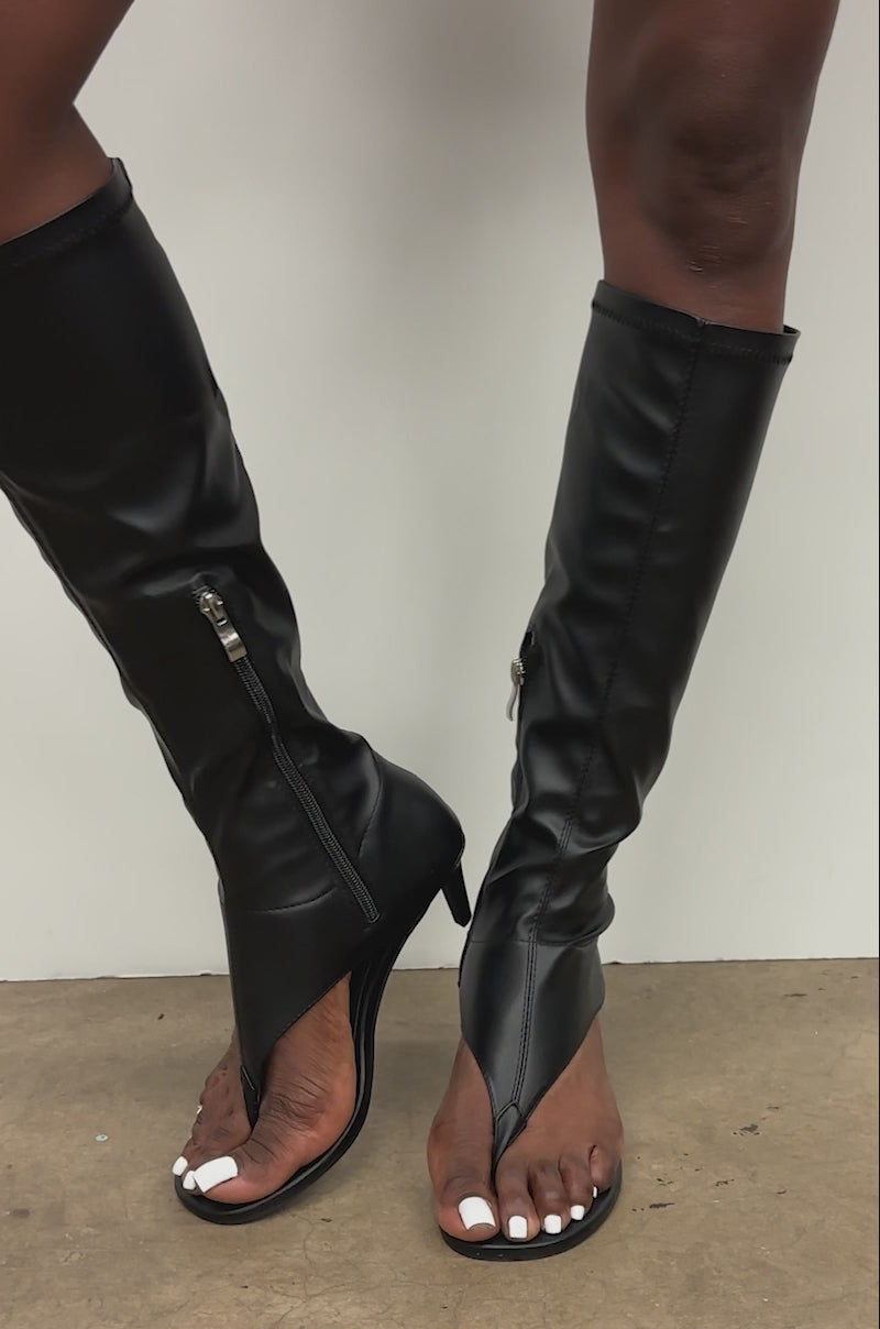 A video of someone wearing the AZALEA WANG CECILE Open Toe Sandal Boot in black stands against a plain light wall, with only their legs visible.