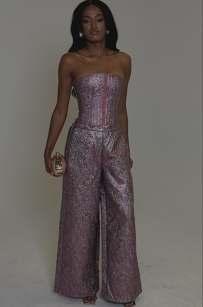 A video of a woman posing in heels against a plain backdrop, wearing NO COMPETITION BROCADE TROUSER—strapless, metallic pink and silver with a textured finish and wide-leg silhouette—paired with a small clutch.