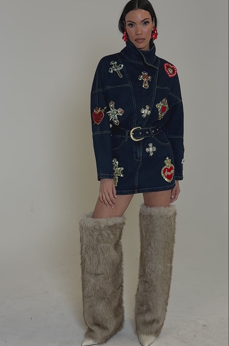 Video of woman modeling the PRETTIEST PROBLEM DENIM PATCHWORK MINI DRESS, featuring a belted waist and jeweled heart and cross details. She completes the look with red earrings, furry knee-high boots, and flowing long hair.