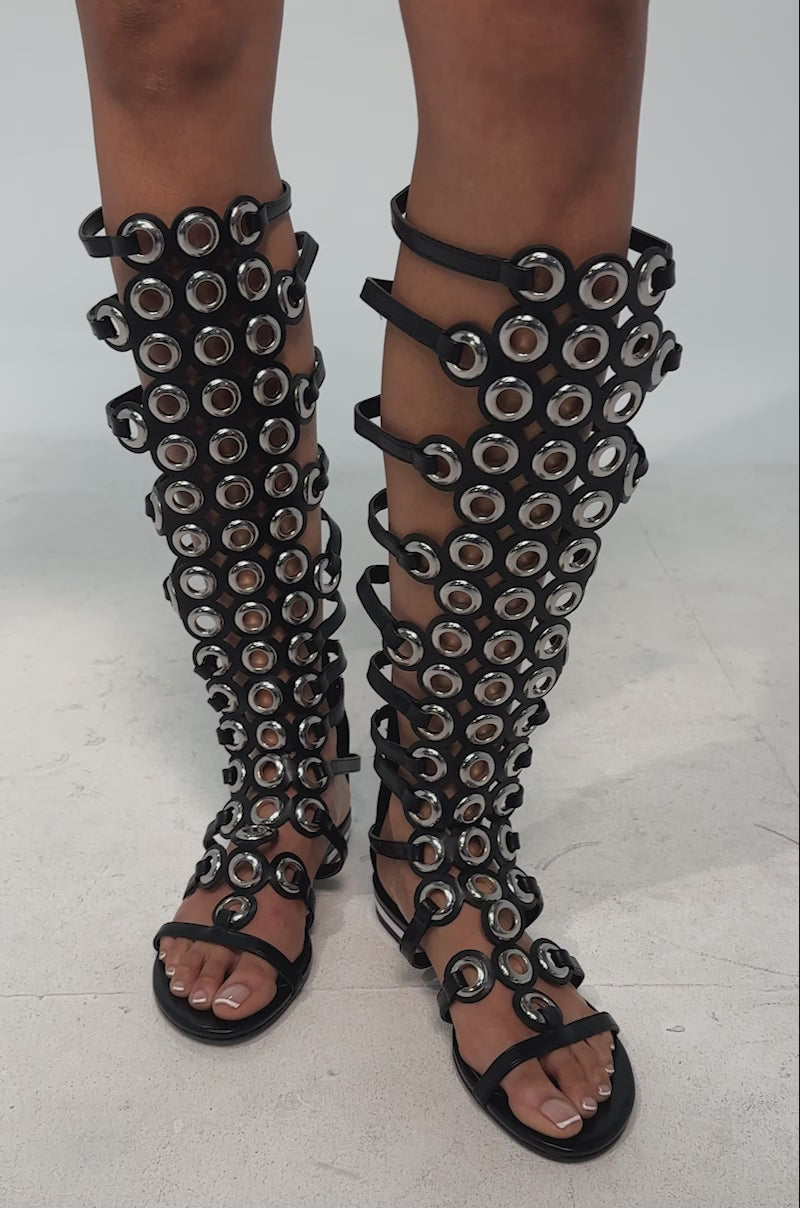 A video of a model wearing the AZALEA WANG LUCE BLACK GROMMET DETAIL FLAT GLADIATOR SANDAL, a knee-high faux leather shoe with multiple straps and metallic eyelet accents.