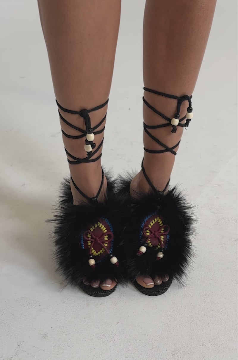 A video of a model wearing the AKIRA GOLD UMIKA BLACK FUR BLOCK SANDAL features black faux fur, colorful embroidery, beaded accents, and lace-up ankle straps for a stylish boho-chic look.