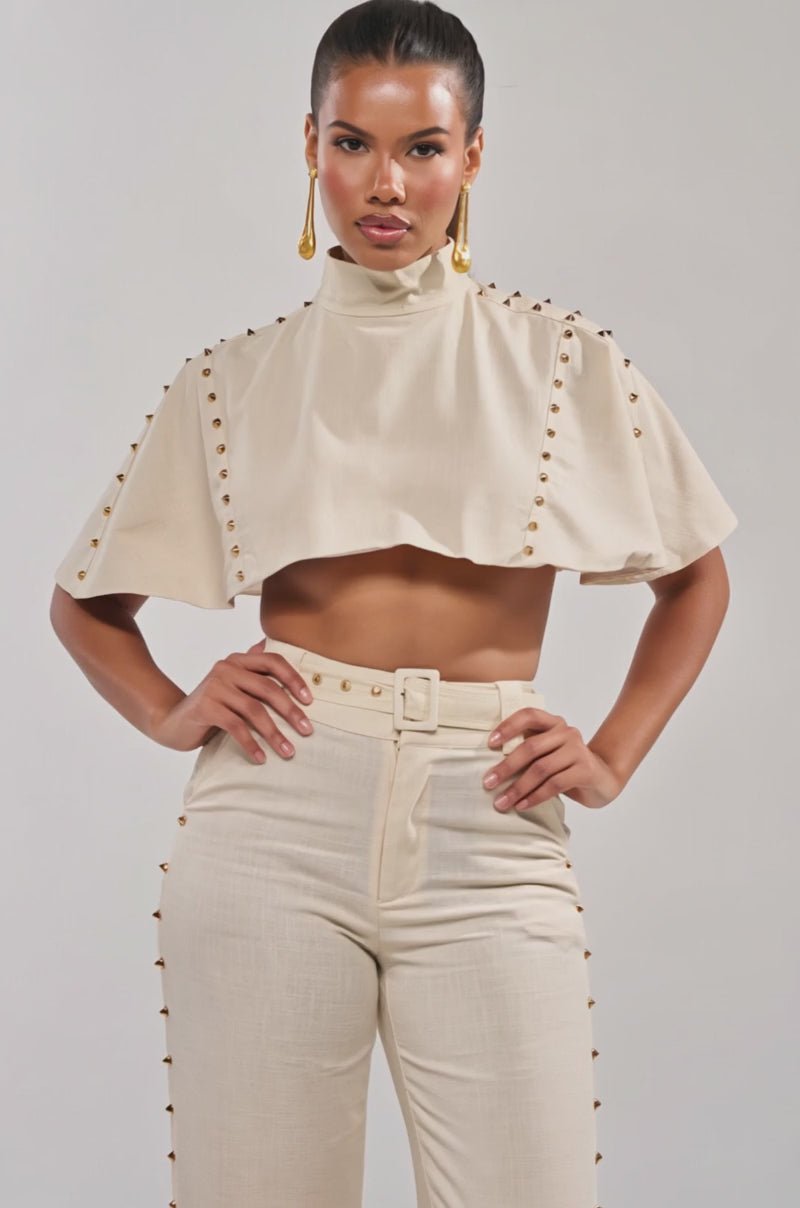 Video of woman modeling the MAEVE LINEN STUDDED BLOUSE—a cream, gold-studded cape top—paired with high-waisted pants and statement gold earrings. Her hair is pulled back, making it perfect for effortless summer style.