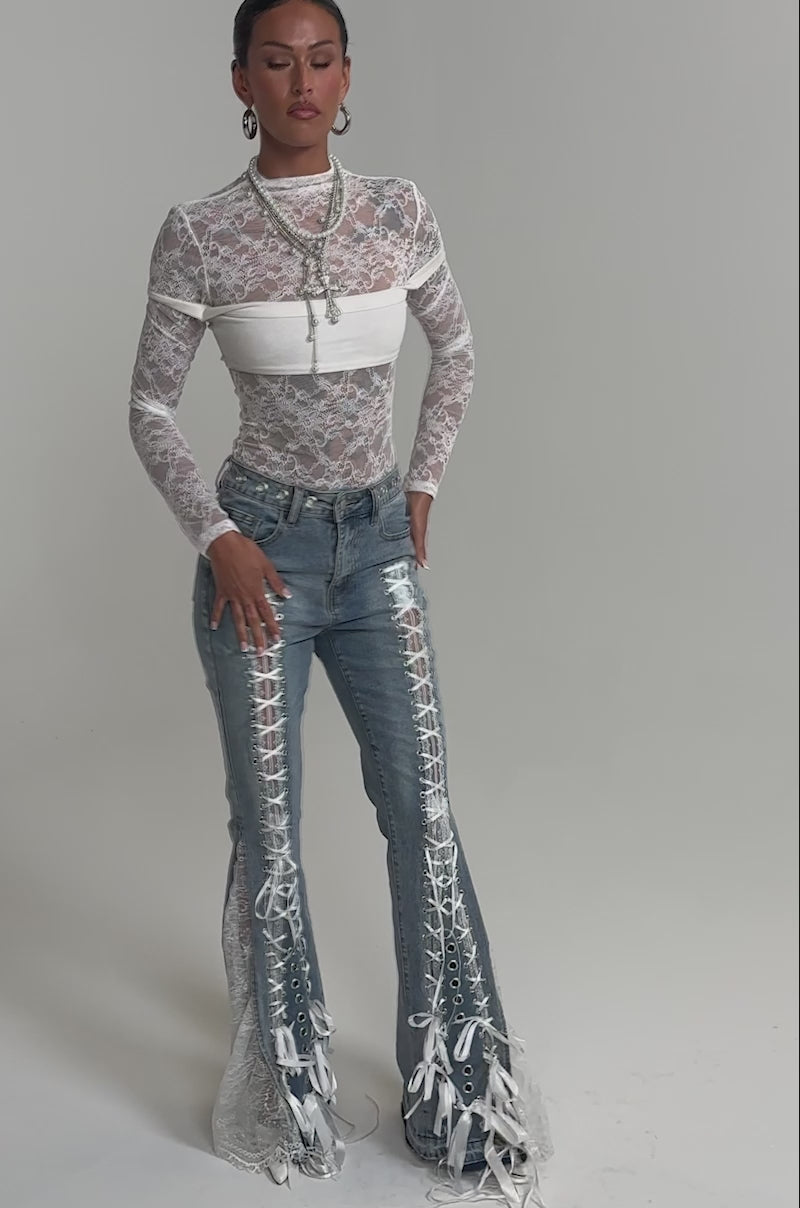 Video of woman modeling the LOVE ME LIKE THAT LACE UP FLARE DENIM—light blue high-rise wide-leg jeans with sheer white lace panels, white ribbon and grommet details—paired with a long-sleeve white lace top.