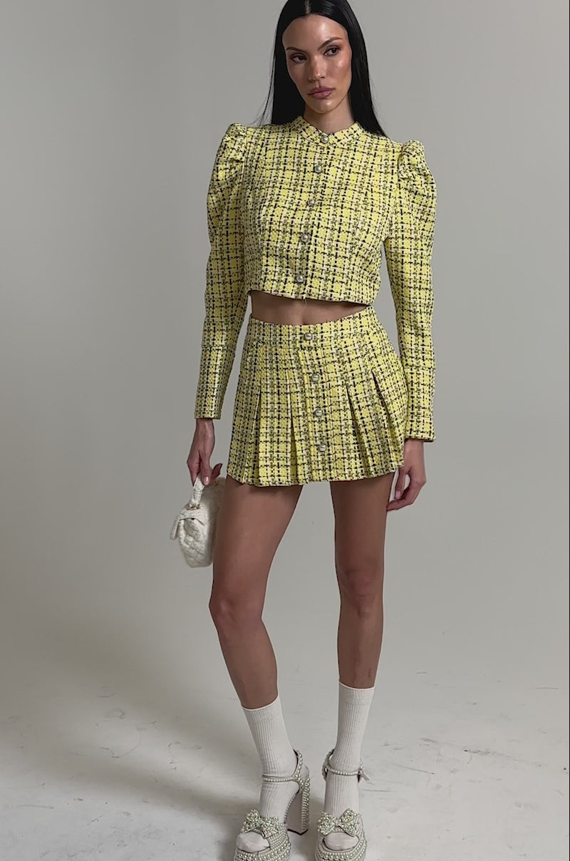 A video of a woman in the FINN TWEED BLAZER, a yellow plaid blazer with pearl buttons, paired with a matching mini skirt, white socks, chunky platform heels, and a small white handbag.