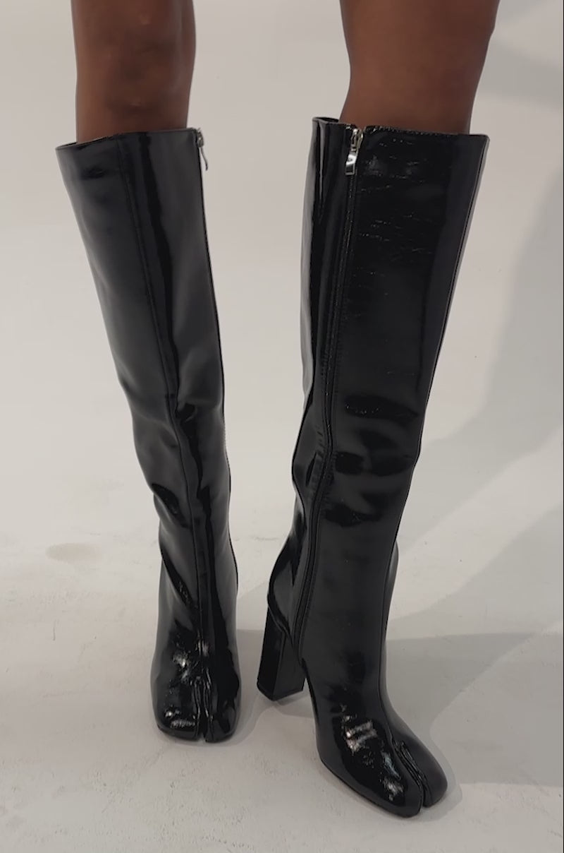 Video of a woman modeling the AZALEA WANG BILHANA BLACK SPLIT TOE BLOCK BOOTS—knee-high, shiny black faux leather footwear. 