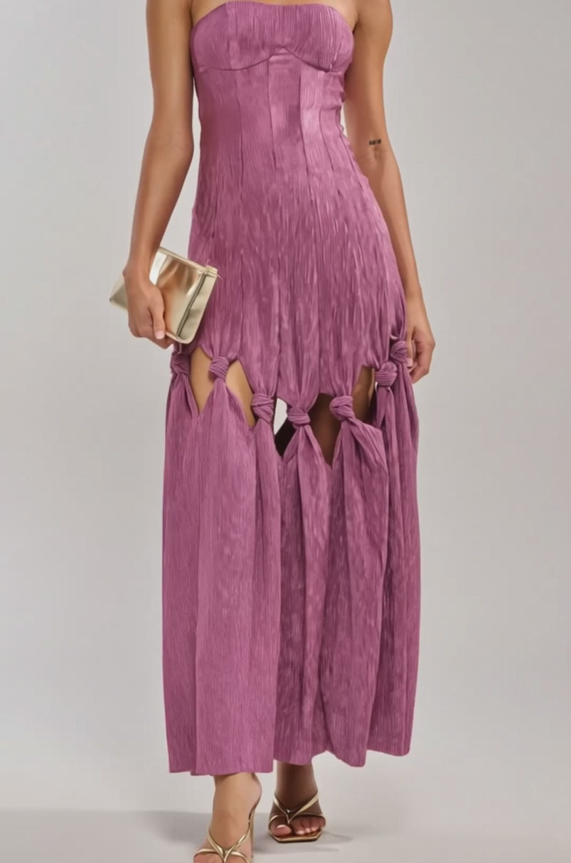 A video of a woman in the BORN READY KNOTTED STRAPLESS MAXI DRESS—mauve, strapless, with knots above the knees—pairs it with gold hoop earrings, gold sandals, and a gold clutch.