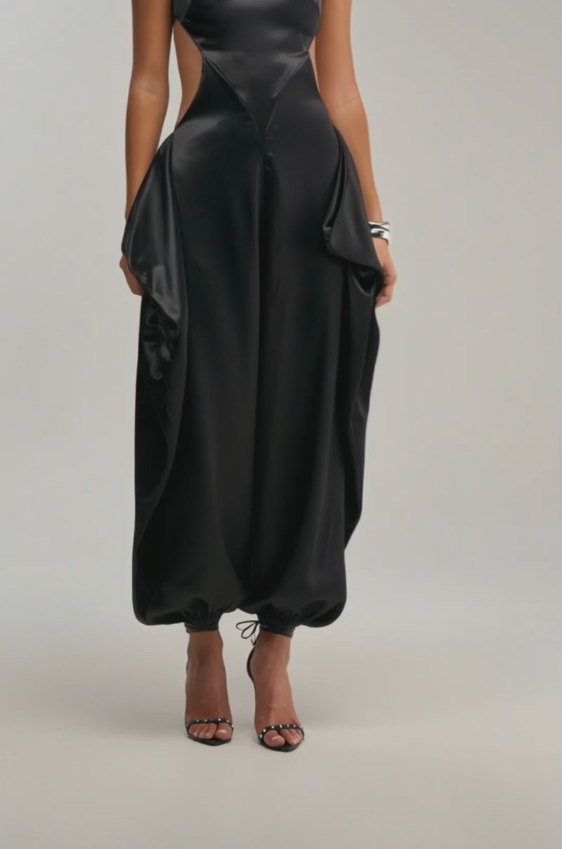 A video of a woman in the A MOMENT IN SATIN JUMPSUIT, featuring black satin with wide draped hip panels and gathered ankles, paired with black open-toe heels.