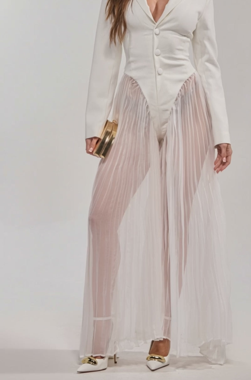 Video of woman modeling the ICON BEHAVIOR PLEATED BLAZER JUMPSUIT, featuring sheer, pleated chiffon pants styled with white high heels with gold accents, set against a plain background.