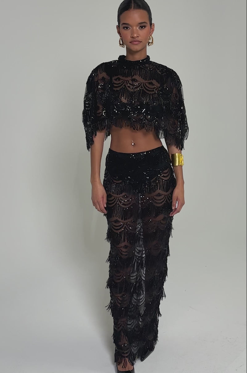 Video of woman modeling the STAR OF THE NIGHT BLOUSE—a sheer black sequin top with scalloped patterns and fringe. She pairs it with gold hoop earrings, a gold cuff bracelet, slicked-back hair, and stands before a light gray background.