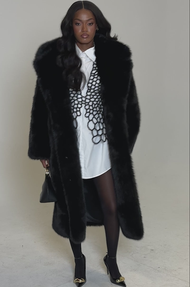 A video of a woman standing against a plain light background wearing the OSLO FAUX FUR COAT IN BLACK over a white button-down shirt with black lace, paired with black tights and black high-heeled shoes with gold tips.