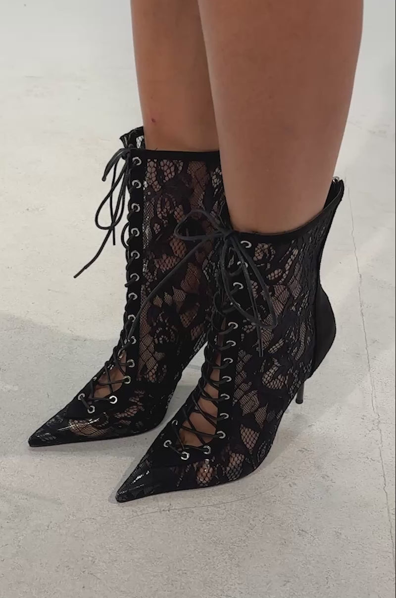 Video of woman modeling the AZALEA WANG VIRTUOSO BLACK LACE STILETTO BOOTIE—black, lace-up, pointed-toe ankle boots with stiletto heels, decorative front laces, a sheer lace overlay, and an elegant high-heeled design.