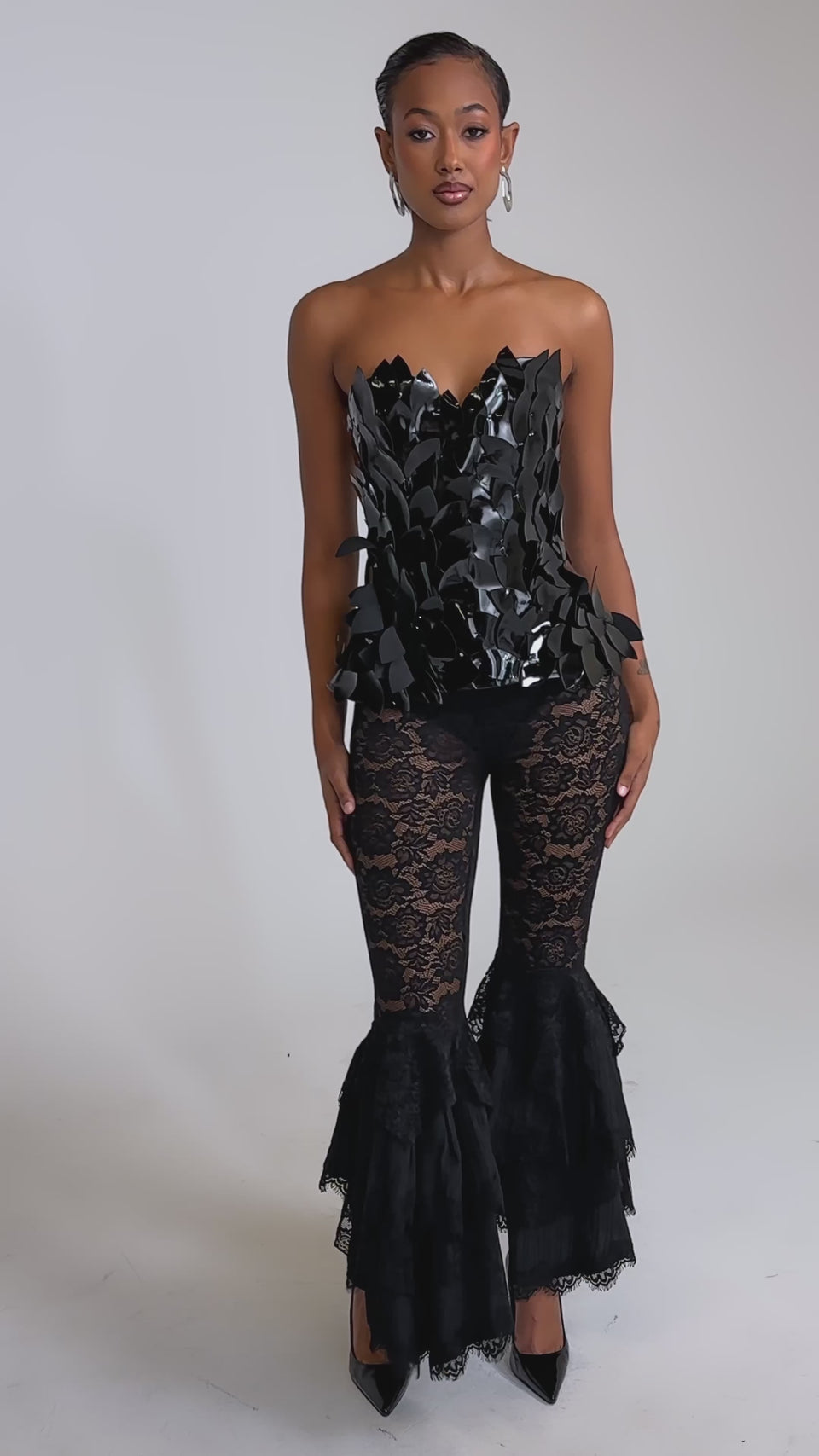 A video of a woman in the DARK NIGHT CORSET, a strapless crop top with leaf-inspired details, styled with black lace bell-bottom pants featuring layered knee ruffles and black pointed heels, set against a plain background.