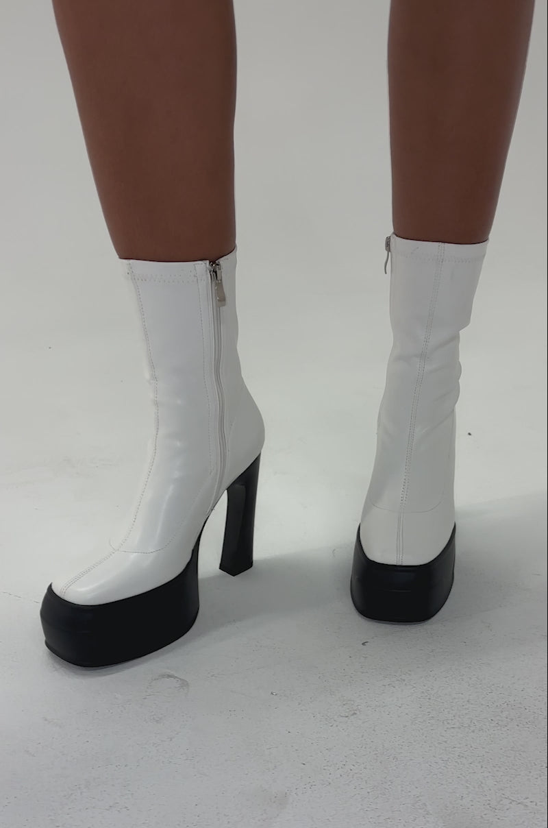 Video of woman modeling the AKIRA GOLD OBSIDIAN WHITE CHUNKY PLATFORM BOOTIE with shiny white faux leather, chunky platform heels, and an ankle zip, standing on a light surface. Only their lower legs and feet are visible.