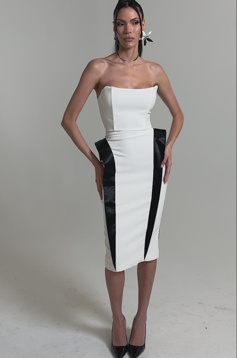 A video of a woman in the CHIC AND SOPHISTICATED STRUCTURED MIDI DRESS IN WHITE with black contrast panels, creating an hourglass silhouette, paired with black pointed-toe heels against a plain light background.