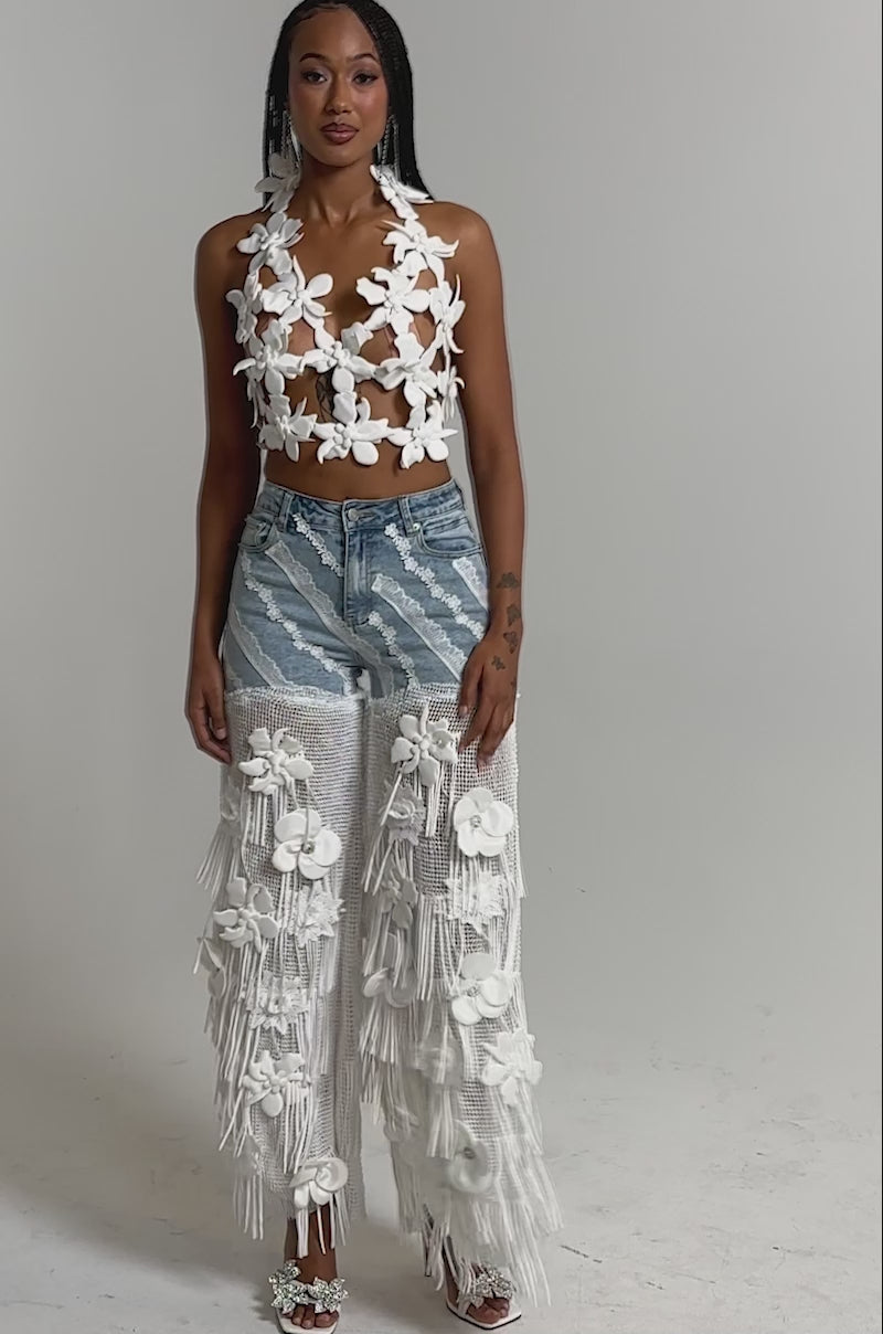 A video of a woman in the FESTIVAL GIRL RELAXED DENIM—crochet fringe pants with sheer, fringed white overlays and 3D flowers—paired with a floral crop top.