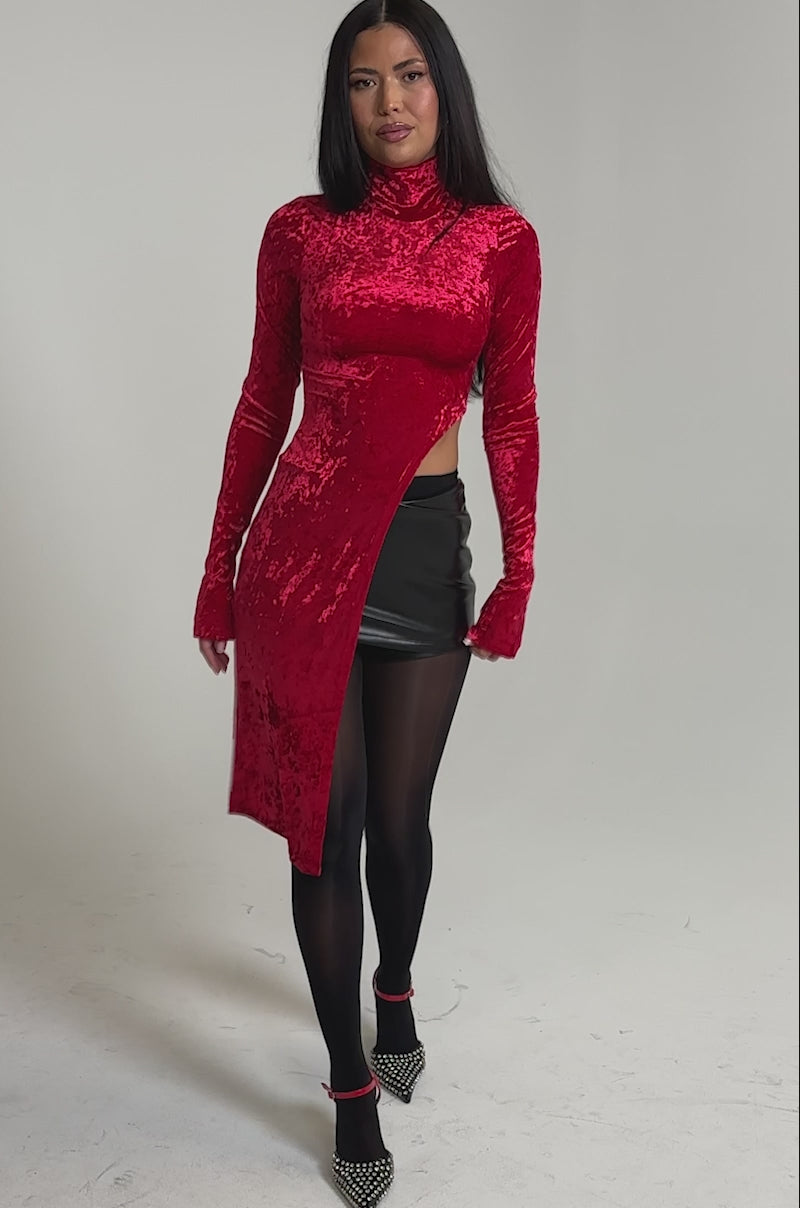 A video of a woman in the LOVE IS IN THE AIR VELVET ASYMMETRICAL LONG SLEEVE BLOUSE, paired with a black leather mini skirt, tights, and silver ankle-strap heels. 