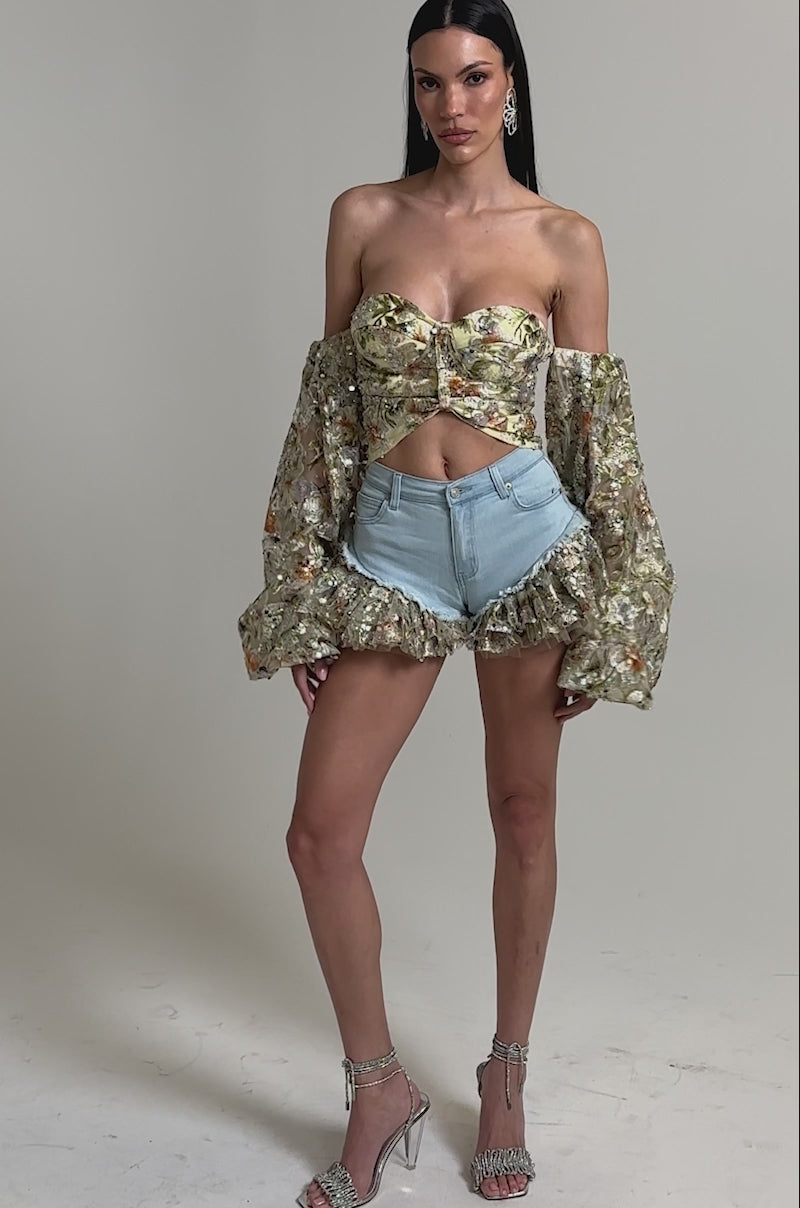 A video of a woman in the QUEEN OF IT ALL FLORAL DENIM SHORT, paired with a floral off-shoulder crop top featuring puffy sleeves, tulle ruffle trim, and silver high-heeled sandals.