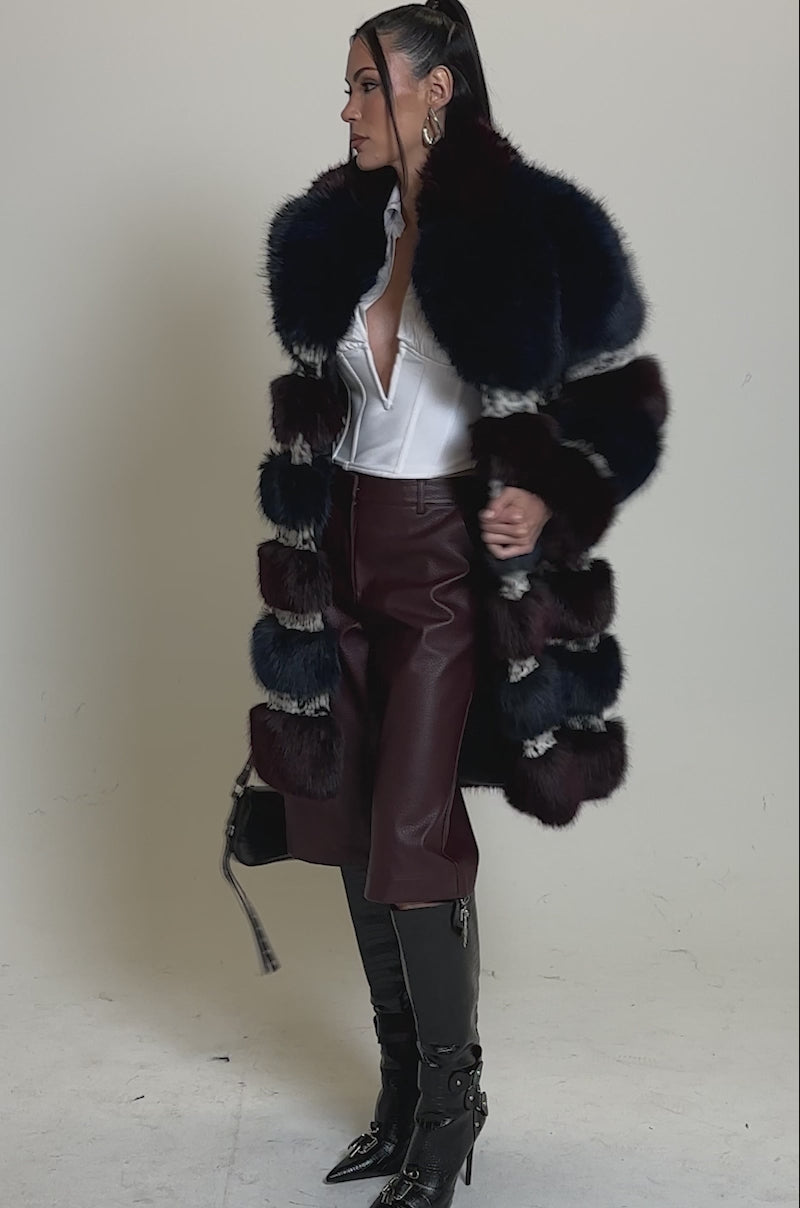 Video of woman modeling the EGO PANELED FAUX FUR COAT featuring horizontal blue, burgundy, and black stripes, styled with knee-high black boots and a black handbag against a neutral background.