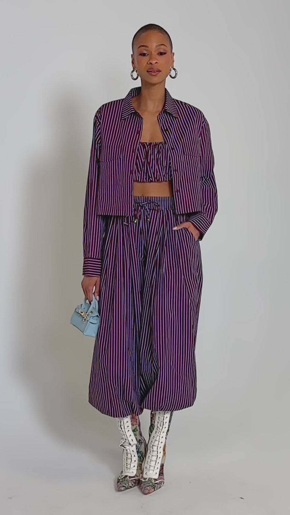 Video of woman modeling the COULD BE DIFF BUTTON DOWN in maroon and blue stripes over a ruffled crop top and high-waisted pants. Large hoop earrings complete her stylish ensemble.