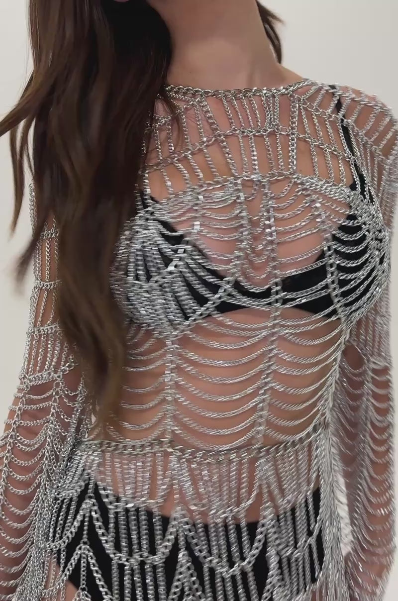 Video of woman modeling the BACKSTAGE PASS METAL BODY CHAIN MINI DRESS over a black bra and high-waisted bottoms, holding a black handbag and standing confidently against a plain gray background.