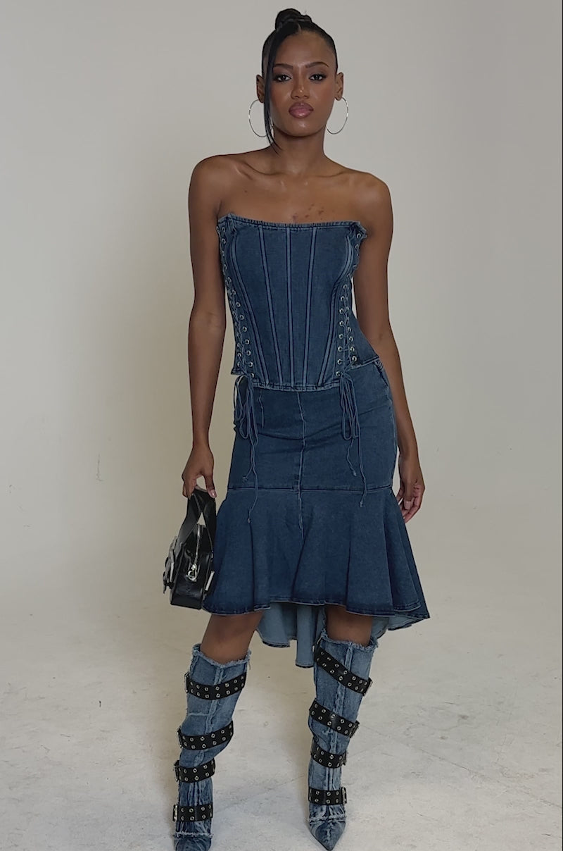 Video of woman modeling the YOU WISH DENIM MIDI SKIRT, a contoured blue denim midi skirt with an asymmetrical ruffled hem, styled with matching knee-high denim boots featuring multiple black buckle straps