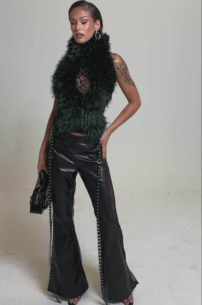 A video of a model posing against a plain light background, a person wears the FEEL ME BABY FUR BLOUSE, sheer black top, black lace-up leather pants, and heels, accessorized with a small black handbag.