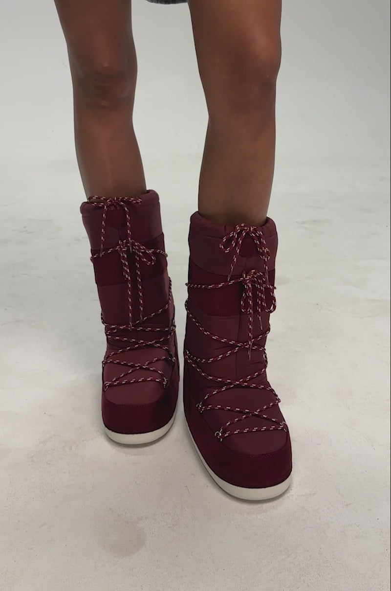 Video of a woman modeling the AZALEA WANG FROSTLING BURGUNDY FLAT BOOT, featuring burgundy moon boots with white wrap-around laces, mid-calf height, and thick rubber soles.