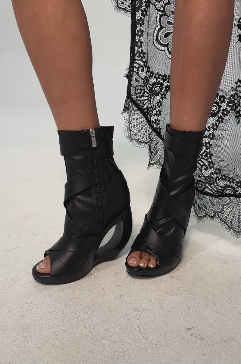 A video of a model wearing the AZALEA WANG IGNITE Black Wedge Open Toe Bootie featuring a belting detail, a striking cutout wedge heel, and overlapping straps.