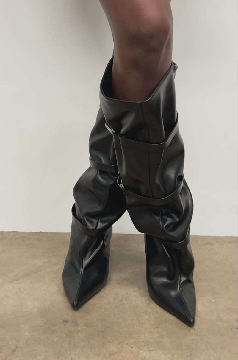 Video of woman modeling the AZALEA WANG KULLERVO Black Silver Clasp Detail Wedge Boots stands against a light wall and concrete floor, with only their legs and part of a black skirt visible.