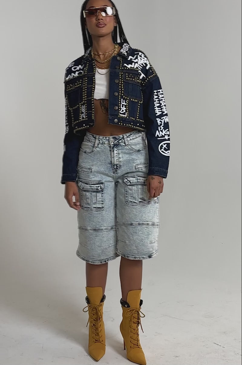 A video of a woman in the RHYTHM OF THE NIGHT DENIM JACKET IN DARK BLUE, graphic tee, light-wash denim cargo shorts, tan lace-up boots, sunglasses, and gold jewelry.
