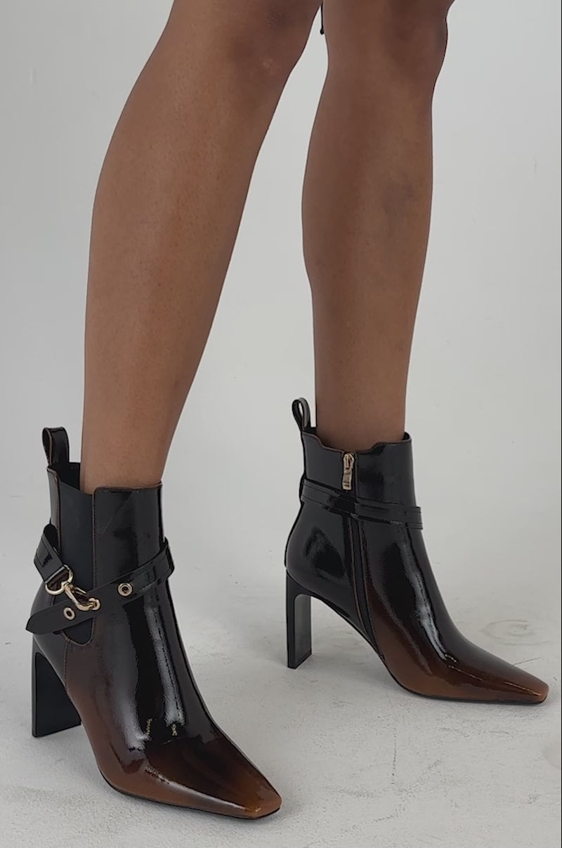 Video of a woman modeling the AZALEA WANG ELYN ESPRESSO BLOCK HEEL BOOTIE—sleek patent leather footwear with gold accents.