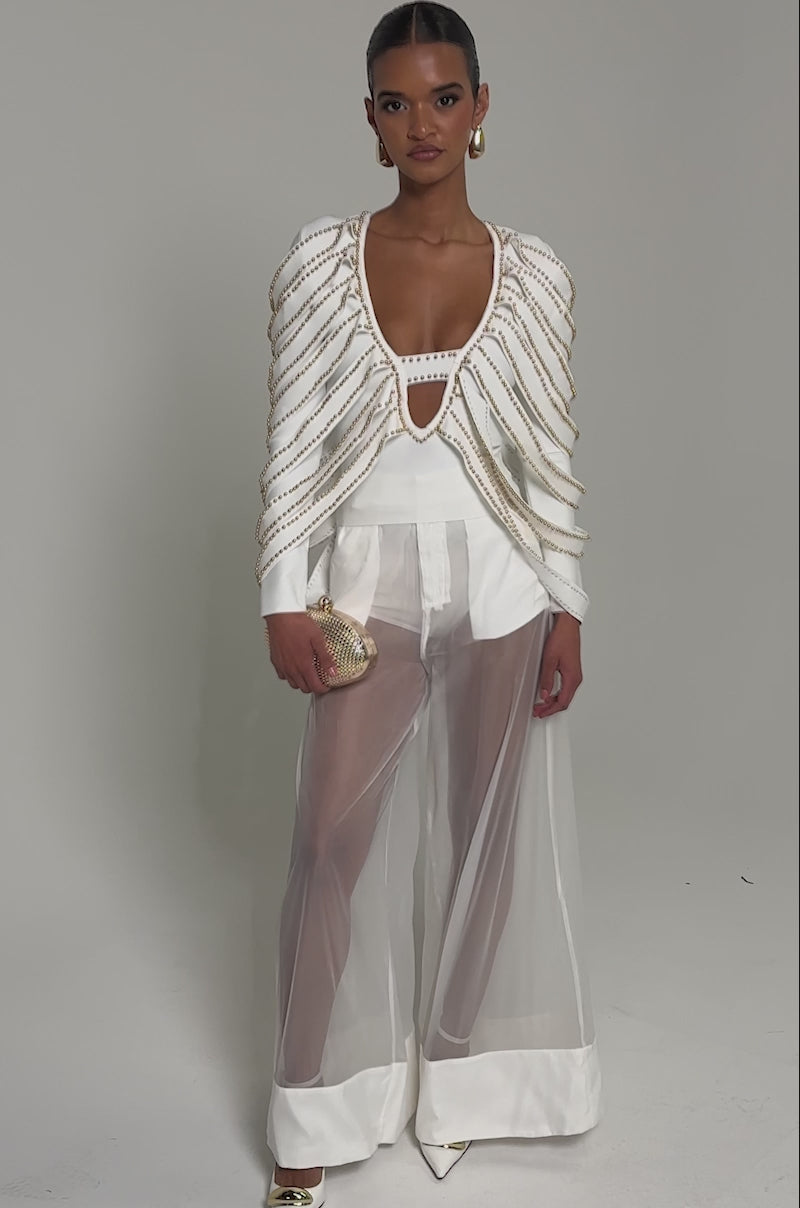 Video of woman modeling the EXTRA AS ALWAYS BLOUSE, featuring a plunging neckline and gold chain details on the shoulders and chest. She completes her chic look with gold hoop earrings, a small gold clutch, and slicked-back hair.