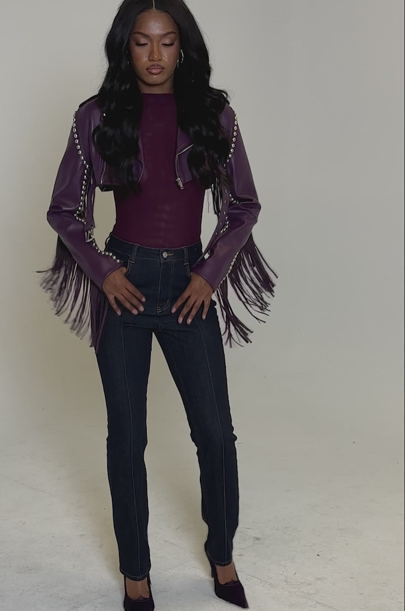 A video of a woman in the HOLD EM CROPPED FRINGE MOTO, a purple cropped jacket with silver hardware, paired with a matching top, dark blue jeans, and purple heels. Her long wavy hair flows against a plain light background.