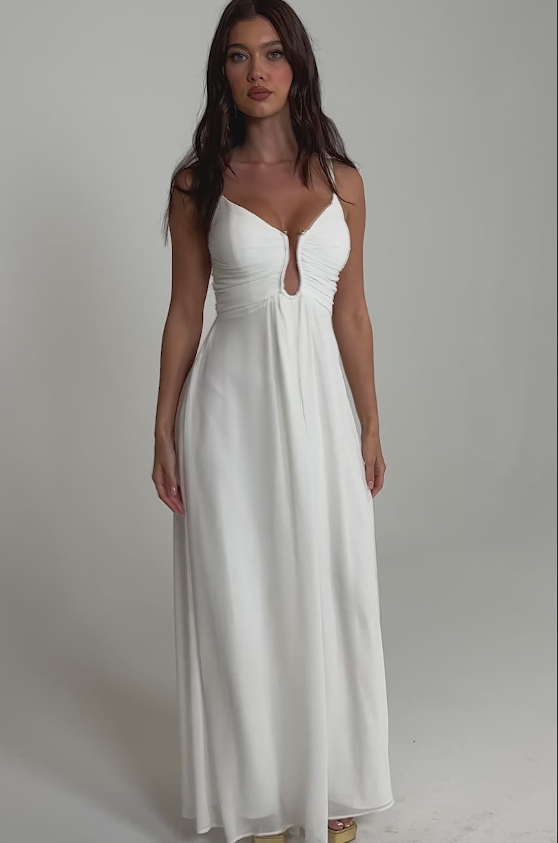 A video of a woman wearing the LATE NIGHT IN KAUAI JUMPSUIT IN WHITE—a sleeveless wide-leg style with deep V-neck and keyhole cutout—paired with gold platform heels.