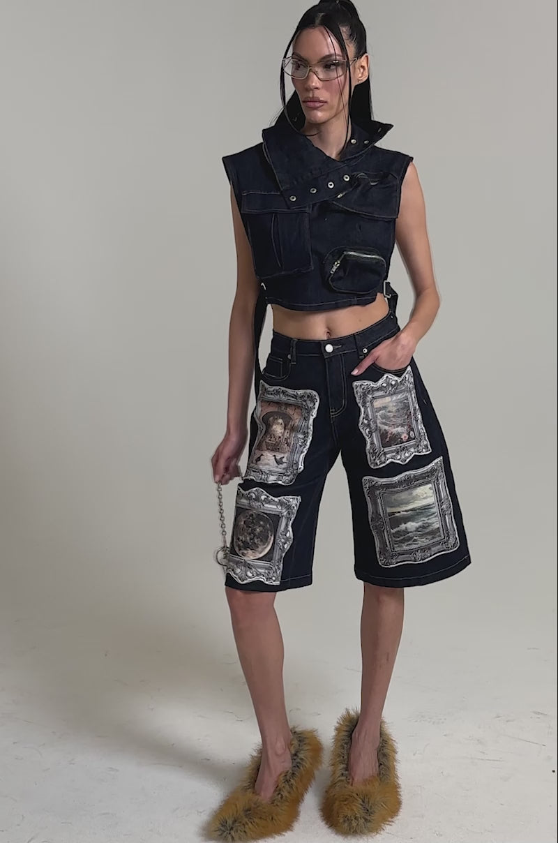 A video of a woman wearing the MODEL BEHAVIOR TOP IN DARK BLUE DENIM, dark denim shorts with printed images, a small cage purse, clear glasses, and fluffy orange slippers stands against a plain light background.
