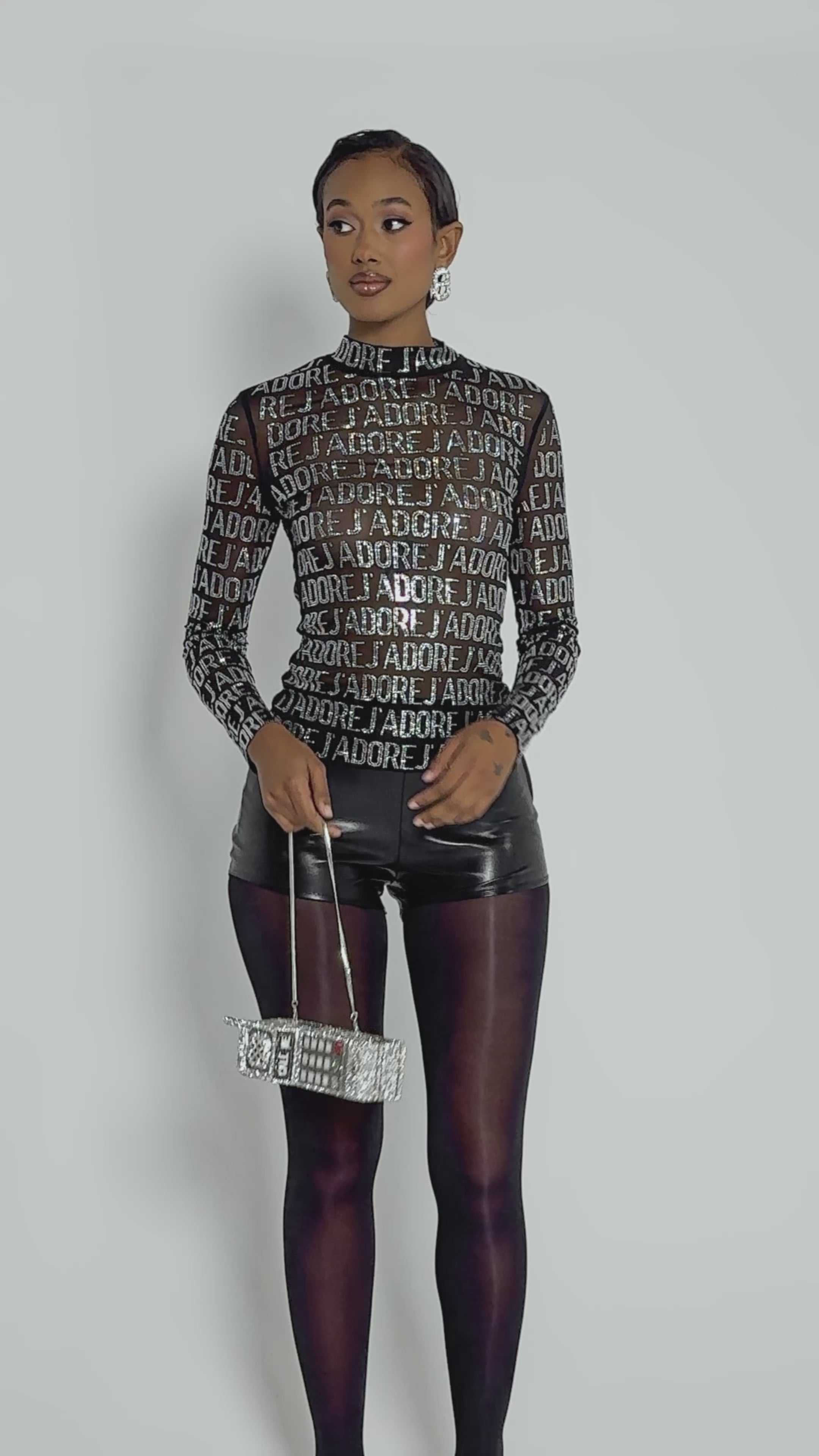 Video of woman modeling the J’ADORE EMBELLISHED MESH TOP with white text, paired with black shorts and sheer black tights.