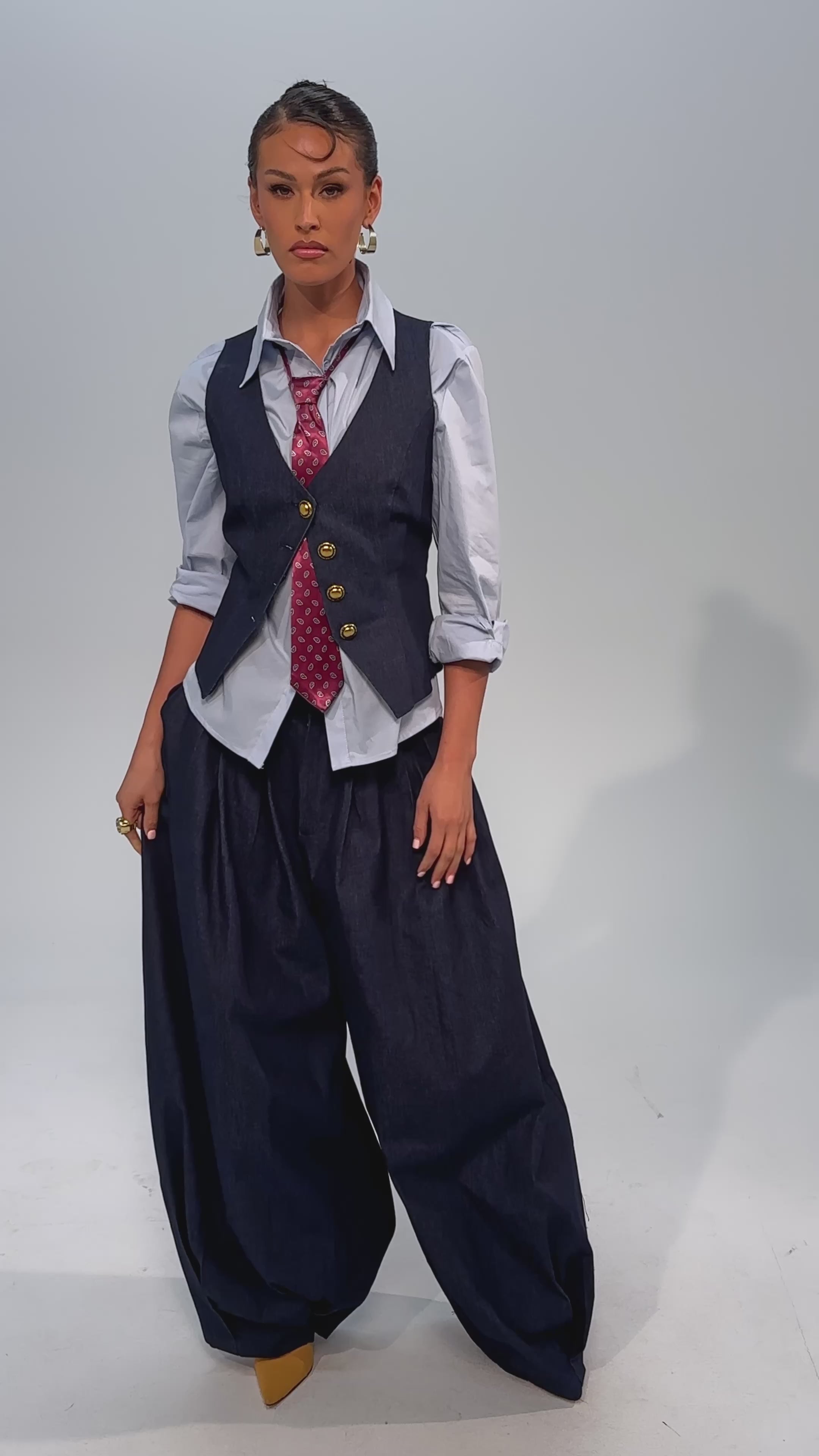 Video of woman modeling the BUSINESS IN THE FRONT TROUSER: dark wash, high-rise, wide-leg denim paired with a matching vest, light blue shirt, red patterned tie, and tan high-heeled lace-up boots.