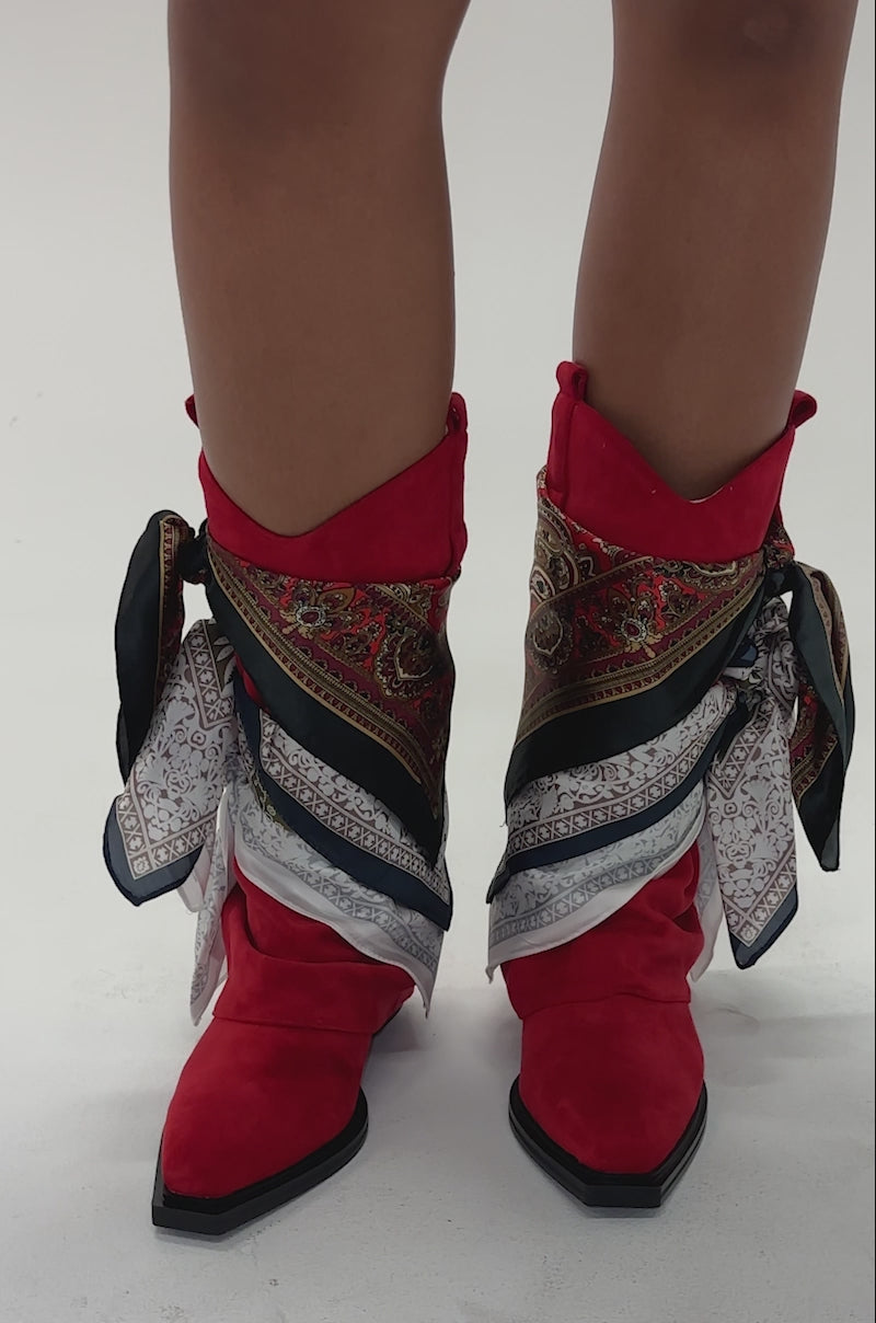 Video of woman modeling the AZALEA WANG COBALT RED FAUX SUEDE SCARF EMBELLISHED WESTERN BOOTIE, a person stands with legs crossed against a light gray background. The booties feature patterned silk scarves tied around the ankles.