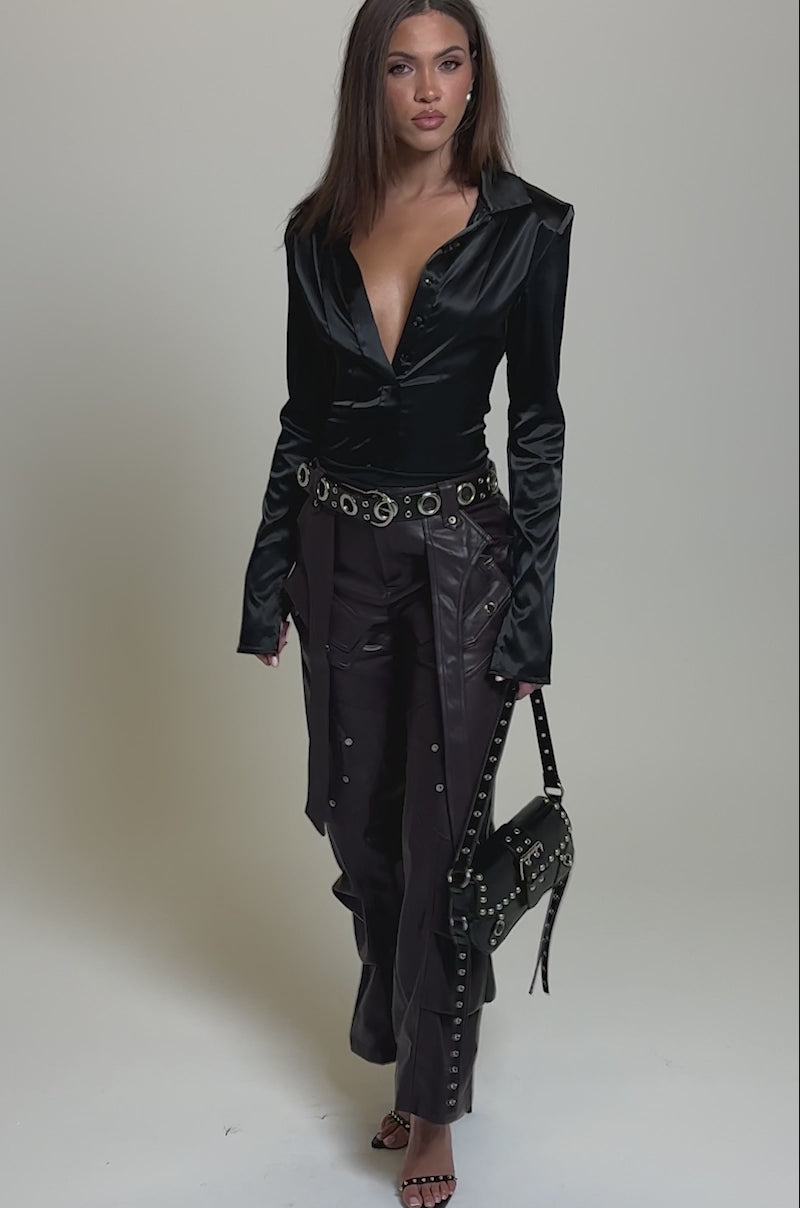 Video of woman modeling the THE BEST PLUM FAUX LEATHER CARGO TROUSER in dark purple with decorative straps and silver rivets, paired with black sandals and a black studded handbag against a plain light gray background.