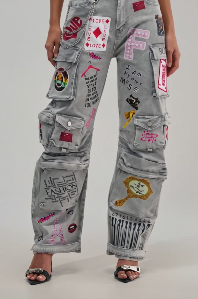 A video of a woman wearing the RAJ CARGO DENIM, featuring colorful patches and playful graphics like hearts and barcodes, styled with open-toe black heels for a bold streetwear look against a plain backdrop.
