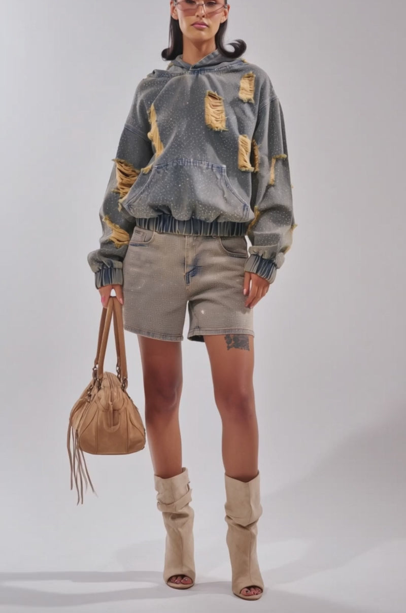 Video of woman modeling the DEMI RHINESTONE DENIM HOODIE and matching jean shorts, slicked-back hair, tinted sunglasses, and a small handbag—an ideal festival streetwear look against a neutral backdrop