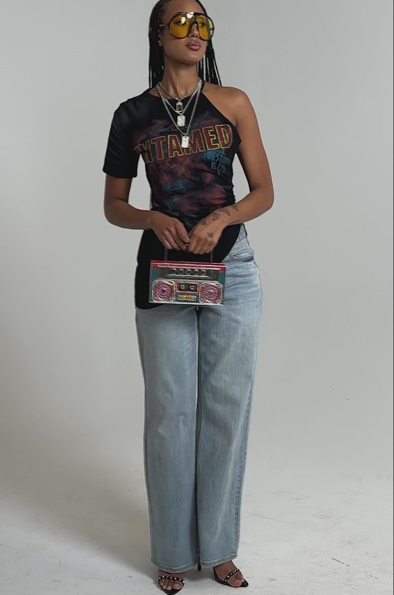A video of a person wearing the UNTAMED SINGLE SLEEVE ASYMMETRICAL TEE, light blue wide-leg jeans, black heels, and holds a colorful rectangular clutch.